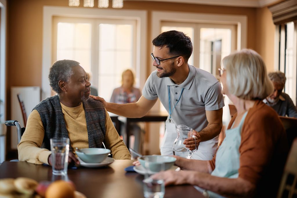 Young healthcare worker talking to seniors during lunch at assisted living facility.